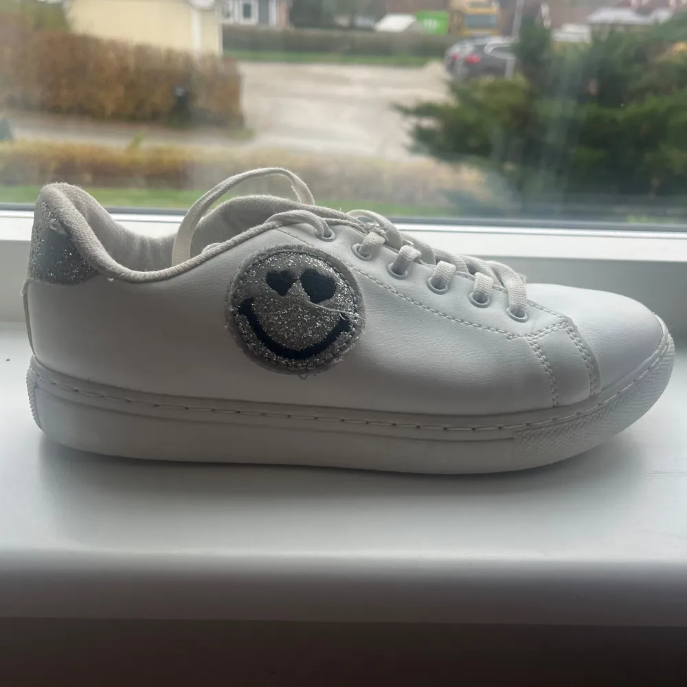 Snygga vita sneakers från SmileyWorld med en glittrig smiley på sidan. Skorna har snörning och en bekväm passform. Köpta för ett år sen men knappt använda. De är i mycket bra skick och redo för ny ägare. Storlek 37. Skor.