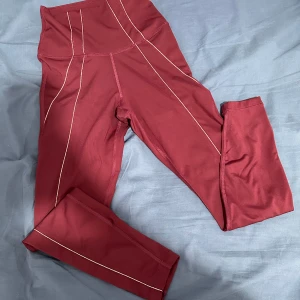 Röda leggings från Nike - Snygga röda leggings från Nike i ett mjukt och stretchigt material. De är högmidjade och har en smickrande passform som är perfekt för träning eller vardagsbruk. Diskreta sömmar och en liten Nike-logga på midjan ger en stilren look. Perfekta för dig som vill ha både komfort och stil!