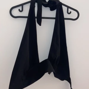 Svart knytbar halterneck topp - Snygg svart halterneck topp med knytband i nacken. Perfekt för sommarens fester eller en kväll ute. Toppen är ärmlös och har en stilren design som passar till det mesta. Ett måste i garderoben för en chic och enkel look!