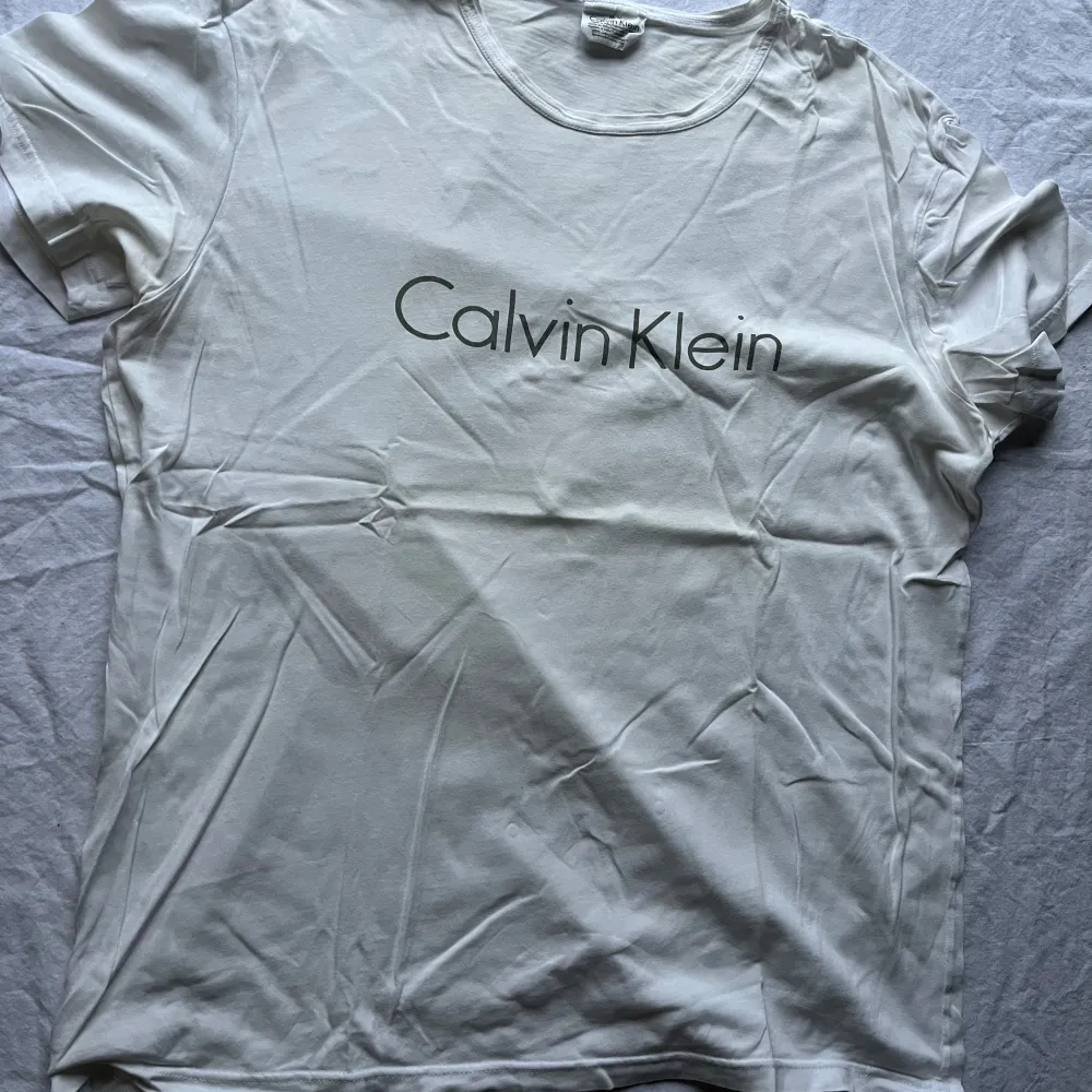 Säljer en klassisk vit t-shirt från Calvin Klein i storlek M. Den är gjord av 100% bomull och har en stilren design med märkesloggan tryckt på framsidan. Perfekt för en avslappnad och stilren look. T-shirten är kortärmad och i bra skick.. T-shirts.