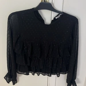 Svart blus från Gina Tricot - Säljer en superfin svart blus från Gina Tricot i storlek 158/164. Den har långa ärmar och är dekorerad med små prickar och volanger som ger en söt och trendig look. Perfekt för både vardag och fest! 🖤