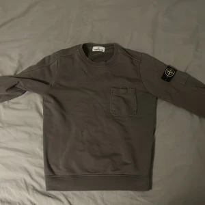 Grå sweatshirt från Stone Island - Säljer en snygg grå sweatshirt från Stone Island. Den har en liten bröstficka och det ikoniska märket på ärmen. Perfekt för en casual look. Tröjan är i bra skick men har lite slitage vid ärmsluten. Passar perfekt till höst och vår. Självklart ÄKTA