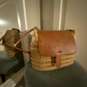 Beige och brun axelväska i flätat material - Säljer en superfin axelväska i flätat beige material med bruna skinn detaljer. Perfekt för sommarens utflykter eller en dag på stan. Väskan har en praktisk storlek och en lång axelrem för bekväm passning. Den stängs med ett lock i brunt skinn.