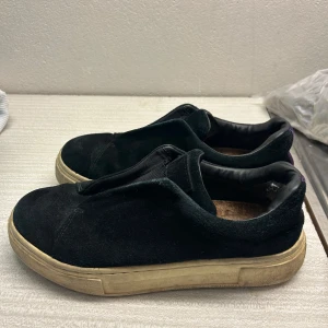 Svarta sneakers i mocka - Säljer ett par svarta sneakers i mocka med en cool lila detalj på hälen. De har en sliten look på sulan och små skavanker i hälen men inget som stör, men är fortfarande i bra skick. Perfekta för en avslappnad stil och funkar året runt. Sköna att ha på sig och lätta att matcha med olika outfits. 