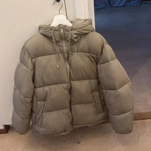 Beige pufferjacka med huva - Säljer en snygg beige pufferjacka i mycket bra skick. Jackan har en praktisk huva och dragkedja framtill. Perfekt för kyliga höst- och vinterdagar. Den är långärmad och har en skön, vadderad känsla. Passar både till vardags och lite mer avslappnade tillfällen.