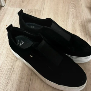 Svarta sneakers från K Cobler - Snygga svarta sneakers från K Cobler i mocka med vit sula. Perfekta för en casual look och passar till nästan allt. De är i bra skick och har en stilren design utan snörning, vilket gör dem enkla att ta på och av. Perfekta för både vardag och fest!