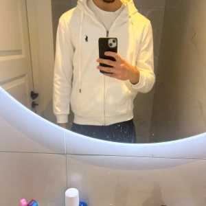 Vit hoodie från Polo Ralph Lauren - Säljer en snygg vit zip up hoodie från Polo Ralph Lauren i storlek L. Den har en dragkedja framtill och en klassisk logga på bröstet. Helt ny oanvänd. Ettiketen sitter kvar. Helt ny från fabriken. Fick den som present men passade tyvärr inte, säljer därför vidare den för billigt. MVH Elt.Wear 