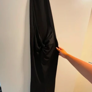 Svart långklänning - Säljer en elegant svart långklänning i syntetmaterial. Klänningen har en stilren design med en knutdetalj framtill som ger en snygg touch. Perfekt för festliga tillfällen eller en utekväll. Passar bra till både sommar och vinter med rätt accessoarer.