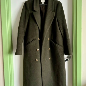 Mörkgrön kappa i ullblandning  - Säljer en stilren mörk olivgrön kappa i ullblandning med dubbelknäppning. Den har långa ärmar och en klassisk krage. Perfekt för höst och vinter, och ger en elegant touch till vilken outfit som helst. Kappan är i mycket bra skick och redo för nya äventyr! 