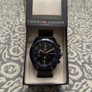 Tommy Hilfiger herrklocka - Säljer en stilren Tommy Hilfiger herrklocka med blå urtavla och svart läderarmband. Klockan har tre subdials och silverfärgade visare med röda detaljer. Perfekt för både vardag och fest. Kommer i originalförpackning. Är också oanvänd.