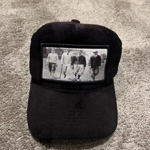 Svart keps med vintagebild - Säljer en svart keps med en cool vintagebild framtill. Perfekt för att ge din outfit en retro touch. Justerbar passform med nät baktill för extra ventilation. Passar både till vardags och fest! Kepsen har tappat några pluppar i spännet men det finns 4 kvar.