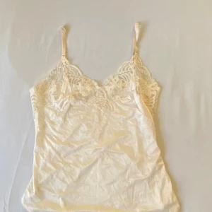 Krämvit linne med spetsdetaljer - Säljer ett supergulligt krämvit linne med spetsdetaljer. Det är ärmlöst med smala axelband och har en romantisk känsla. Perfekt för sommardagar eller som ett sött lager under en kofta. Materialet är mjukt och bekvämt i silkesmatrial, vilket gör det idealiskt för både vardag och fest.