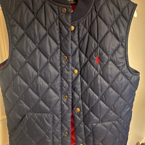 Mörkblå quiltad väst från Polo Ralph Lauren - Snygg mörkblå quiltad väst från Polo Ralph Lauren med röd insida. Västen har knappar framtill och en broderad logga på bröstet. Perfekt för höst och vår! Väldigt lite använd och är i bra skick!