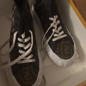 Fendi - Säljer ett par snygga sneakers från Fendi i mycket bra skick. De har ett coolt mönster i brunt och svart med vita snören och sulor. Perfekta för att ge din outfit en lyxig touch. Kommer med originalförpackning och dustbag samt kvitto. Nypris ca 7000 kr