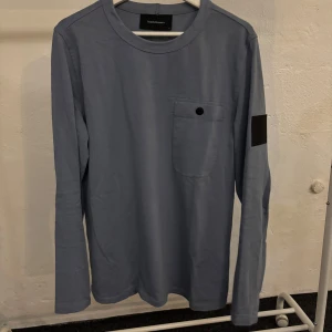 Blå långärmad tröja från Peakpreformance - Säljer en stilren blå långärmad tröja från Peak preformance. Den har en praktisk bröstficka med knapp och en svart patch på ärmen. Perfekt för både vardag och lite mer avslappnade.