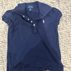 Mörkblå pikétröja från Ralph Lauren - Säljer en snygg mörkblå pikétröja från Ralph Lauren. Den har korta ärmar och en klassisk krage med knappar. Perfekt för en avslappnad men stilren look. Den ikoniska loggan syns på bröstet. Passar både till vardags och lite finare tillfällen.