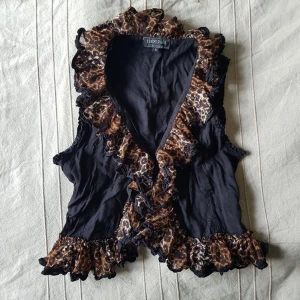 Vintage topp med volanger, retro - Stängning med 3 knäppen på framsidan och snöre där bak för att justera bredden. I fint använt skick.  Anmärkningar: knäppet högst upp sitter löst så den är snabblagad med en säkerhetsnål, men är lätt att sy tillbaks. - se sista bild.