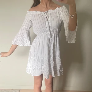 Vit randig offshoulder klänning - Säljer en superfin vit offshoulder klänning med tunna ränder. Den har trekvartsärmar och en söt knytning framtill. Perfekt för sommarens alla fester eller en mysig dag på stan. Klänningen är i mycket bra skick och har en luftig passform som är både bekväm och stilren. Senast använd var på midsommar och blev en stor favorit bland mitt umgänge🤍