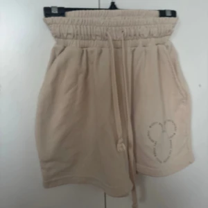 Beige shorts med Mickey Mouse-tryck - Säljer ett par superbekväma beige shorts med Mickey Mouse-tryck på sidan. De har en elastisk midja med dragsko för perfekt passform. I mycket bra skick, inga skavanker!