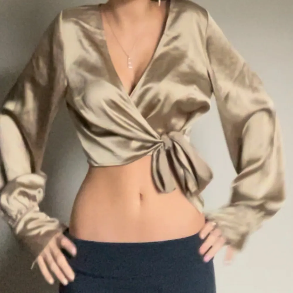 Snygg beige omlottopp i satin med långa ärmar och knytning i midjan. Perfekt för en festlig kväll eller en stilren vardagslook. Toppen har en v-ringad design som ger en elegant touch. Passar bra till både jeans och kjol!. Toppar.