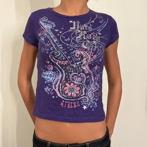 Lila t-shirt från Hard Rock Cafe - Cool lila t-shirt från Hard Rock Cafe med ett stort gitarrtryck och texten 'Athens'. Perfekt för en avslappnad stil eller konsert. T-shirten är kortärmad och har en tight passform. Den är i bra skick!💜