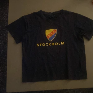 Mörkblå t-shirt med DIF Stockholm tryck - Säljer en mörkblå t-shirt med DIF Stockholm tryck i gult och rött. Perfekt för alla Djurgården-fans! T-shirten är kortärmad och i bra skick. Passar perfekt till vardags eller matchdagar.