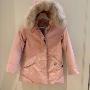 Rosa woolrichjacka med pälsdetaljer - Säljer en superfin rosa woolrich parkajacka med fluffig päls på huvan. Jackan har långa ärmar och stängs med både dragkedja och knappar. Perfekt för kyliga dagar och ger en mysig känsla. Passar perfekt till vintern och hösten!