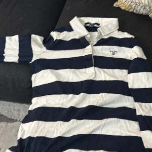 Randig pikétröja från GANT - Säljer en snygg randig pikétröja från GANT i blått och vitt. Tröjan har en klassisk krage och knappar framtill. Perfekt för en avslappnad men stilren look. Passar bra till både vardag och lite finare tillfällen. 🌀