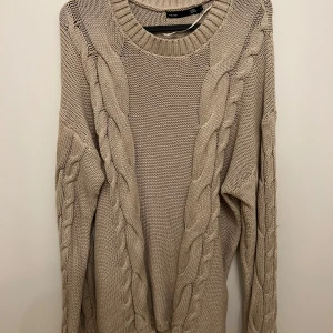 Beige stickad tröja från lager 157 - Säljer en supermysig beige stickad tröja från lager 157. Den har ett snyggt flätmönster på framsidan och är perfekt för kyliga dagar. Tröjan är långärmad och har en rund halsringning. Perfekt för höst och vinter!