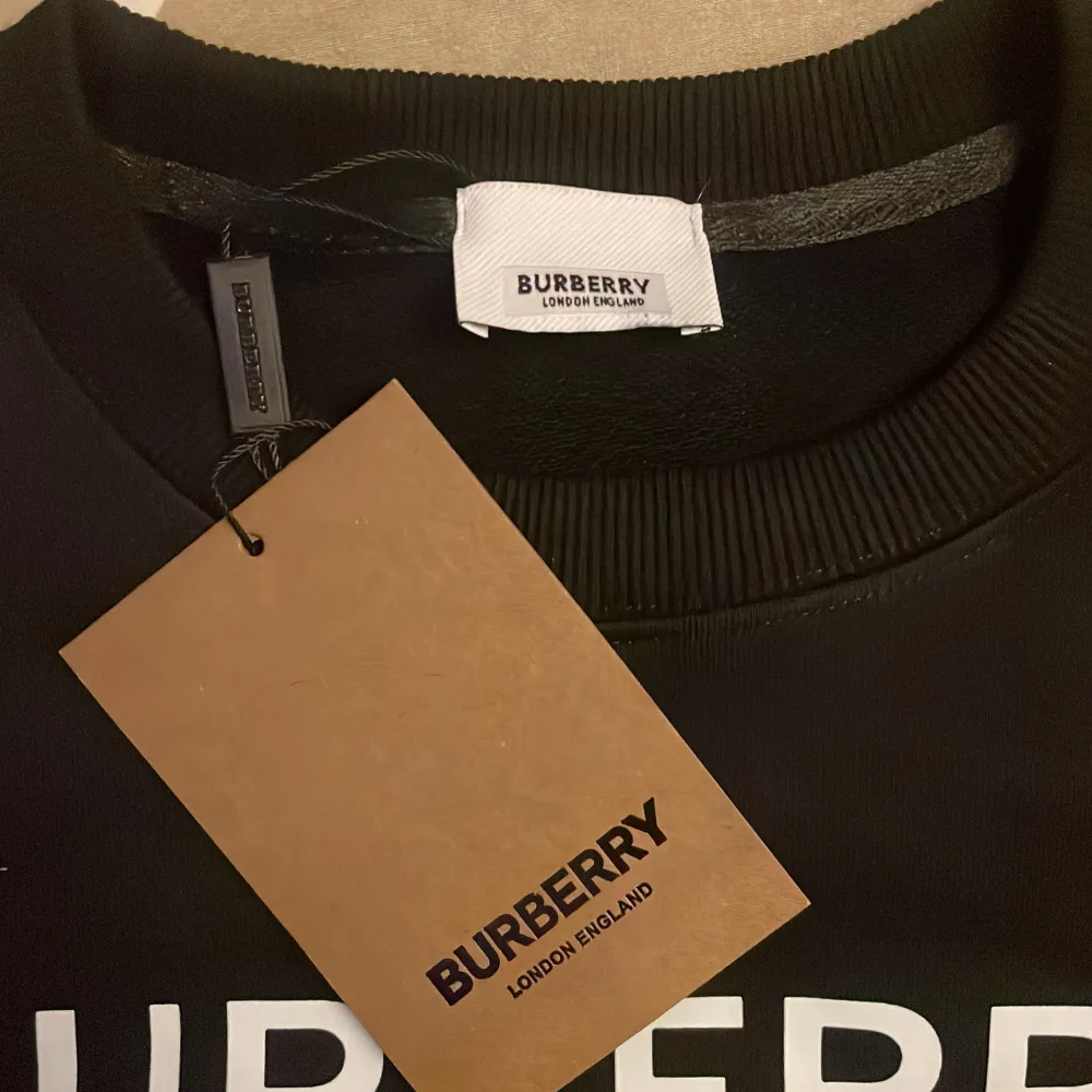 Säljer en snygg svart tröja från Burberry med tryck i vitt och brunt. Tröjan har en klassisk rund hals och är perfekt för både vardag och fest. Den är i nytt skick och har en stilren design med texten 'Burberry' och 'London' tryckt på framsidan.. Tröjor & Koftor.