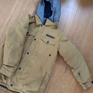 Beige jacka från Burton - Säljer en snygg jacka från Burton i storlek XL. Jackan har en avtagbar huva i grått och flera praktiska fickor med knappar. Perfekt för kyliga dagar med sin varma och tåliga design. Passar både till vardags och utomhusaktiviteter. Stl 160-170 och har room to grow som är ett system Burton har så man kan växa i deras plagg. 
