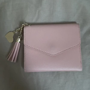 Rosa plånbok med tofsdetalj - Säljer en söt rosa plånbok i syntetmaterial med en charmig tofs och hjärtdetalj i metall. Plånboken har flera fack för kort och en knappstängning. Perfekt för att hålla ordning på dina kort och kontanter. 💖