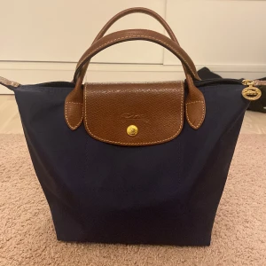 Blå och brun handväska från Longchamp - Säljer en snygg och klassisk handväska från Longchamp i blått och brunt. Väskan har ett stilrent utseende med bruna skinnhandtag och detaljer. Perfekt för både vardag och fest! 👜✨kan prata om priset☺️☺️