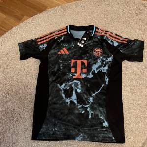 Säljer en ny FC Bayern München fotbollströja i svart med orange detaljer. Tröjan har korta ärmar och ett coolt mönster i svart och grått. Den är märkt med Adidas Aeroready-teknologi för optimal komfort. Perfekt för alla Bayern-fans! 🖤🧡