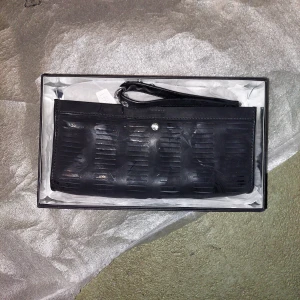 Svart kuvertväska med Swarovski-element - Snygg svart kuvertväska i skinn med coolt mönster och en glittrig detalj från Swarovski. Perfekt för en festkväll! Väskan har en praktisk handledsrem och kommer i originalförpackning.