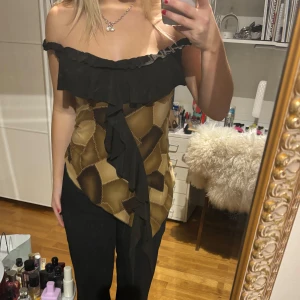 Offshoulder topp med volang - Säljer en snygg offshoulder topp med volangdetaljer. Toppen har ett unikt mönster i bruna och beige toner, perfekt för en utekväll eller fest. Den är i bra skick och ger en chic look med sin asymmetriska design framtill.