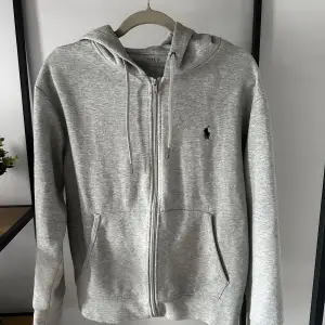 Säljer en stilren grå hoodie från Polo Ralph Lauren i mycket bra skick. Den har en dragkedja framtill och en klassisk logga på bröstet. Perfekt för både höst och vår, med en skön passform och huva för extra mysfaktor. Passar till det mesta i garderoben!