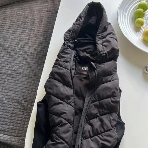 Svart dunväst från Zara - Säljer en svart dunväst från Zara i mycket bra skick. Västen har en dragkedja framtill och en mysig huva med dragsko. Perfekt för höst och vår när du vill hålla dig varm men ändå se stilren ut. Passar till både vardag och lite mer avslappnade tillfällen.