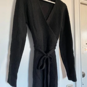 Svart omlottklänning från Nelly - Snygg svart omlottklänning från Nelly i medium. Den har en v-ringad design och ett knytband i midjan för en smickrande passform. Perfekt för både vardag och festliga tillfällen. Klänningen är långärmad och gjord i ett mjukt material som känns skönt mot huden.