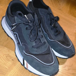 BOSS JONAH RUNN - Sneakers - black one Stl. 43 - Snygga svarta sneakers från Boss (Modell JONAH RUNN - Sneakers - black one)  i NYTT SKICK. Nypris ligger på 2 245 kr ifrån Zalando men jag säljer dom för endast 1099 kr! De har en stilren design med vita och bruna detaljer samt snörning. Perfekta för både vardag och festliga tillfällen. Skorna är inte använda mer än testade. Säljer skorna pga att jag fick dom i present och skorna är för stora för mig har storlek 43.