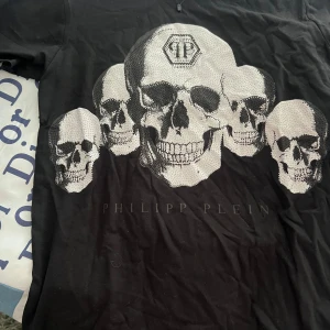 Svart t-shirt med dödskallemotiv från Philipp Plein - Säljer en cool svart t-shirt från Philipp Plein med ett stort dödskallemotiv i silver och svart. T-shirten har en normal passform och är perfekt för en edgy look. Den är i mycket bra skick och passar perfekt till både vardag och fest.