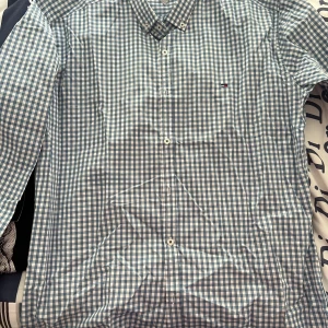 Rutig skjorta från Tommy Hilfiger - Säljer en snygg rutig skjorta från Tommy Hilfiger i storlek XXL. Skjortan är blå och vit med klassiskt rutmönster och har långa ärmar. Perfekt för både vardag och fest! Den är i mycket bra skick och har knappar framtill. Passar perfekt till jeans eller chinos för en stilren look.