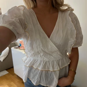 Vit omlottblus med volanger - Superfin vit omlottblus med volanger, puffärmar och mörkgula detaljer från Selected Femme i storlek 38. Skrynklig på bilden men i jättefint skick! 🤍