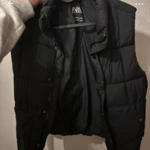 Svart dunväst från Zara - Säljer en svart dunväst från Zara i mycket bra skick. Västen har en stilren design med dragkedja och knappar framtill. Perfekt för höst och vår när du vill hålla dig varm utan att bära en jacka. Passar bra till både vardag och fest.