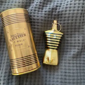 Säljer en lyxig parfymflaska från Jean Paul Gaultier, Le Male Elixir. Flaskan har en cool design i form av en manlig torso i guld och svart. Den kommer i en matchande guldig metallförpackning. Perfekt för den som vill ha en elegant och maskulin doft.