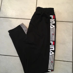 Svarta mjukisbyxor från Fila - Säljer ett par svarta mjukisbyxor från Fila med elastisk midja och logotyp längs sidorna. Perfekta för en avslappnad stil eller träning. De är i bra skick och superbekväma. Passar perfekt för både chill och aktivitet!