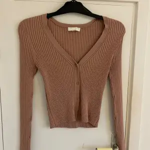 Säljer en superfin beige/rosa ribbad kofta från H&M. Den har en v-ringad design med knappar framtill och är croppad för en modern look. Perfekt för både höst och vår! 💗