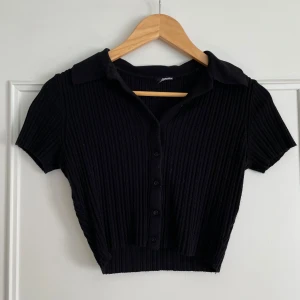 Svart ribbad kortärmad topp - Säljer en snygg svart ribbad topp med korta ärmar och knappar framtill. Den har en klassisk krage och är croppad, vilket gör den perfekt för både vardag och fest. Passar bra till jeans eller kjol för en stilren look.