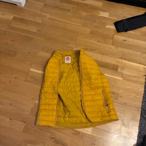 Gul dunväst från Komblen Originals - Säljer en snygg gul dunväst från Komblen Originals. Perfekt för höst och vår, med dragkedja framtill och quiltad design. Västen är i mycket bra skick och passar både till vardags och lite mer avslappnade tillfällen. Ett måste för garderoben om du gillar färg!