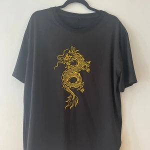 Svart t-shirt med draktryck - Cool svart t-shirt med ett gult draktryck på framsidan. Perfekt för en avslappnad stil eller en utekväll. T-shirten har en normal passform och är kortärmad, vilket gör den idealisk för sommardagar eller som en lager-på-lager-stil.